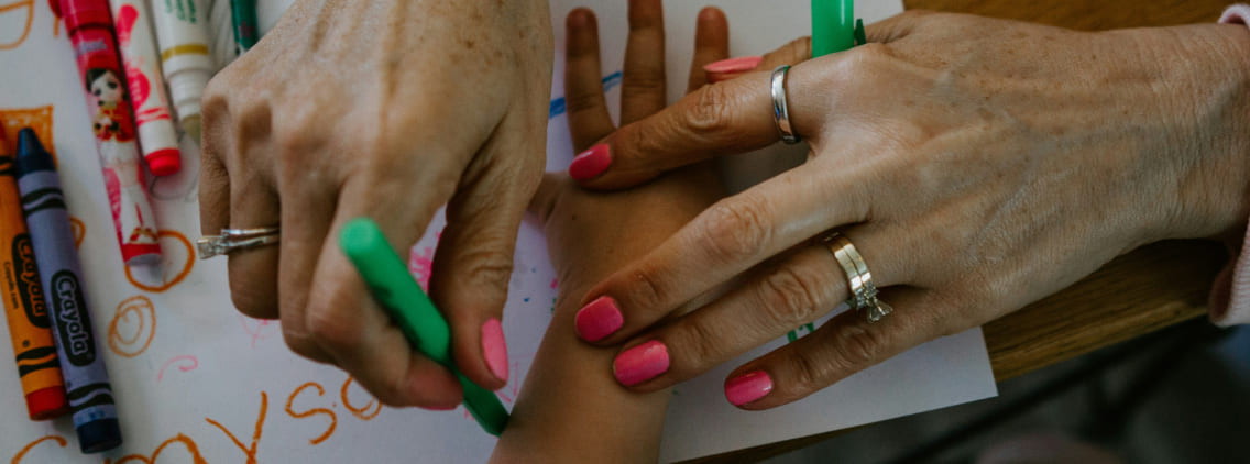A close-up of an adult's hands guiding a child's hand as they draw with markers on a sheet of paper, symbolizing learning, collaboration, and mentorship in a creative setting.
