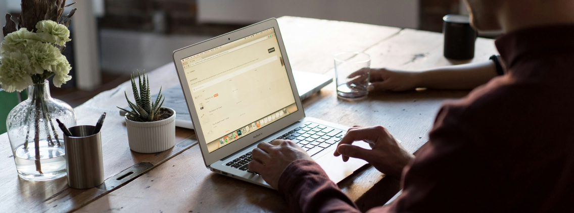 Person working on a laptop in a cozy workspace with a glass of water, potted plants, and office supplies on a wooden table, representing a self-service business intelligence setting