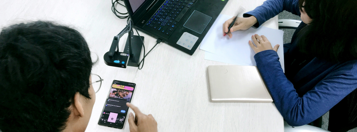 Two people collaborating at a desk—one using a smartphone and the other taking notes on paper, with a laptop and tablet also visible.