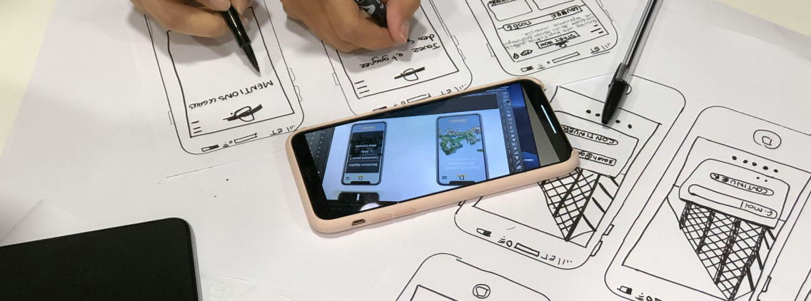 Person drawing wireframes of a mobile app on paper, with a smartphone displaying digital mockups placed on top of the sketches