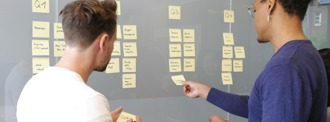 wo men collaborating in front of a glass wall covered with sticky notes organized by quarterly goals, planning tasks using the Kanban method in an Agile work environment