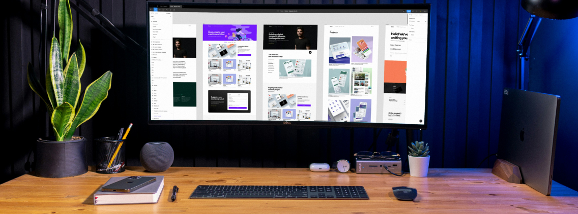Modern workspace setup with an ultra-wide monitor displaying a design portfolio, a wooden desk with a keyboard, mouse, and various desk accessories, illuminated by a stylish desk lamp