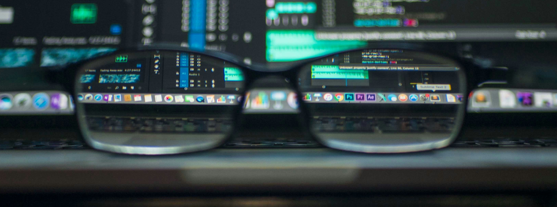 A close-up shot of a pair of glasses resting on a laptop keyboard, with the laptop screen displaying lines of code and digital audio editing software. The focus is on the lenses, which clearly magnify parts of the screen, while the surrounding area remains slightly blurred. The image conveys themes of programming, data analysis, and digital content creation.