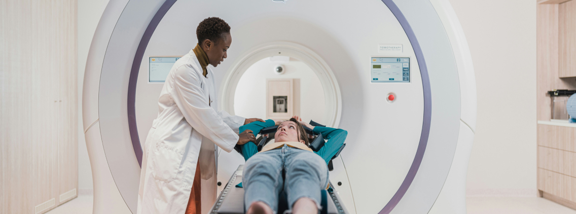 A doctor in a white coat assists a patient lying on a sliding table before entering an MRI machine in a modern medical facility. The patient looks up while the doctor provides reassurance.