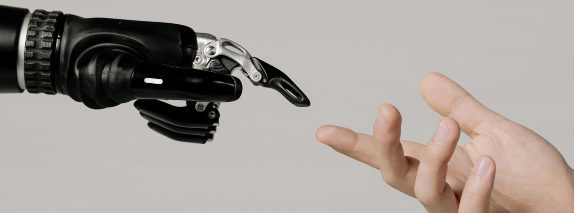 A close-up shot of a robotic hand reaching out to touch a human hand, symbolizing the connection between technology and humanity. The black mechanical prosthetic contrasts with the natural human fingers, emphasizing themes of artificial intelligence, robotics, and human-machine collaboration. The neutral background keeps the focus on the interaction.