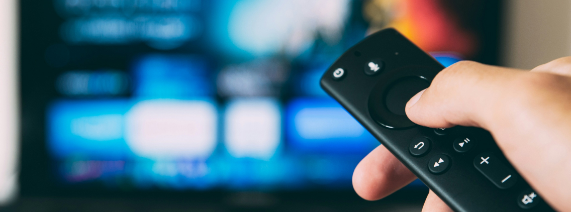 A close-up shot of a person holding a smart TV remote control, navigating a streaming service interface displayed on a blurred television screen in the background. The remote features voice command and navigation buttons, emphasizing modern entertainment technology and digital content consumption.