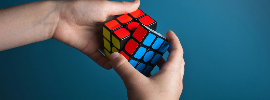 A close-up of a person’s hands solving a Rubik’s Cube against a solid blue background. The cube's vibrant red, blue, and yellow colors contrast with the background, symbolizing problem-solving, logic, and critical thinking. The image represents mental challenges, strategy, and cognitive skills.